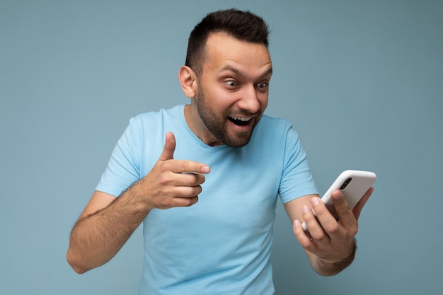 Surpreendido, jovem e bonito moreno barbudo usando uma camiseta azul comum isolada sobre o azul