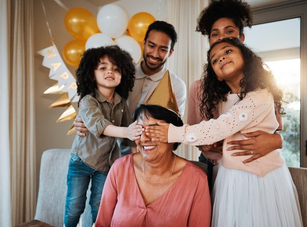 Surpresa de aniversário e filhos com a avó em festa na sala de estar para celebração de evento e confraternização Família comemora e olhos fechados da avó com crianças felizes para presentes em casa