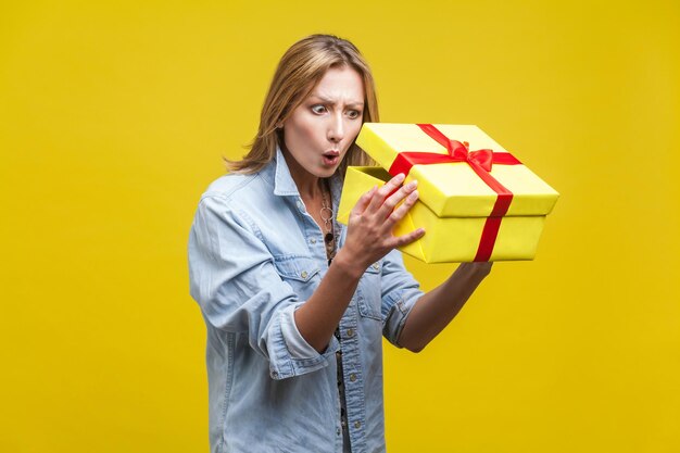 Surpresa de férias Retrato de mulher curiosa atônita em camisa jeans, olhando para dentro da caixa de presente, verificando o que está dentro de unboxing com uau expressão espantada studio shot isolado em fundo amarelo