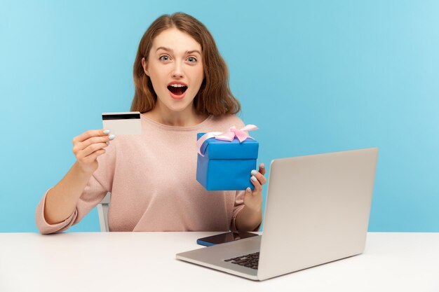 Surpresa excitada linda mulher sentada na mesa com o laptop e segurando a caixa de presente, mostrando o cartão de crédito e olhando para a câmera com espanto, expressão chocada. tiro de estúdio interno, fundo azul