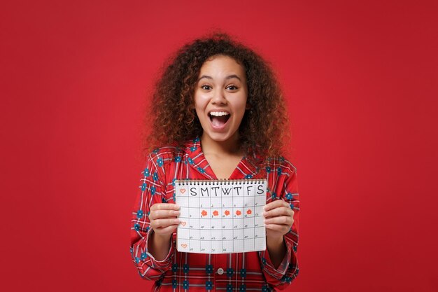 Foto surpresa jovem afro-americana em homewear de pijama descansando em casa isolada em fundo vermelho. relaxe o conceito de estilo de vida de bom humor. segure o calendário de períodos femininos para verificar os dias de menstruação.