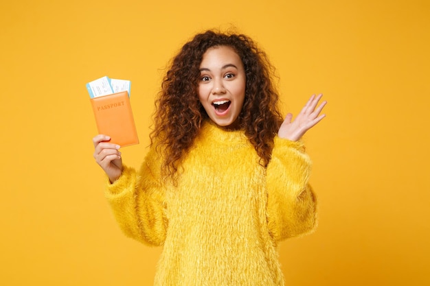 Surpresa jovem afro-americana em suéter de pele posando isolado no fundo da parede amarela laranja. conceito de estilo de vida de pessoas. mock up espaço de cópia. segure os bilhetes de embarque do passaporte, espalhando a mão.