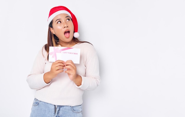 Surpresa jovem asiática de 20 anos no chapéu de Natal recebe um cartão de cupom de vale-presente isolado no fundo branco do estúdio Feliz Natal Conceito