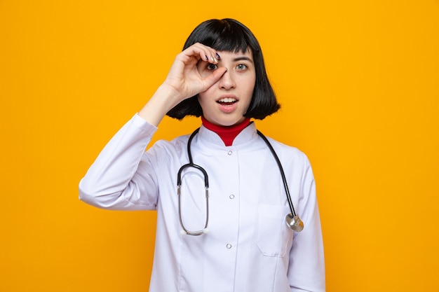 Surpresa, jovem, bonita, caucasiana, com uniforme de médico e estetoscópio olhando por entre os dedos