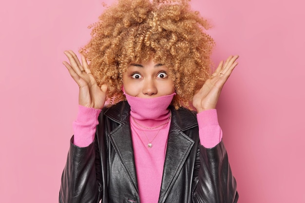 Surpresa jovem excitada com cabelo espesso encaracolado mantém as palmas das mãos levantadas cobre a boca com gola de jumper vestida com jaqueta de couro isolada sobre fundo rosa Conceito de pessoas e reações