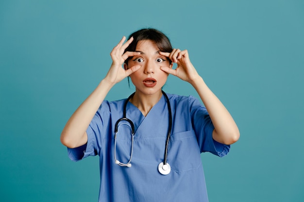 Surpresa mostrando gesto de máscara jovem médica usando estetoscópio uniforme de quinto isolado em fundo azul