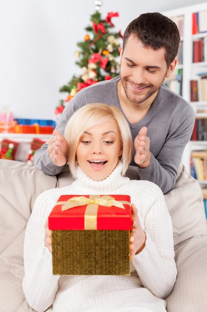 Surpresa! mulher jovem e bonita sentada no sofá segurando uma caixa de presente enquanto o namorado está atrás dela sorrindo