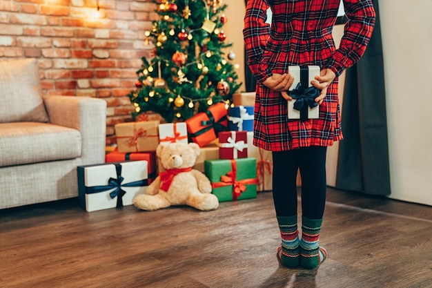 Surpresa para presente de natal em casa decorada perto da árvore de natal. Menina segurando a caixa de presente nas costas. criança vestindo vestido vermelho e meias coloridas em pé no chão de madeira, procurando ursinho de pelúcia.