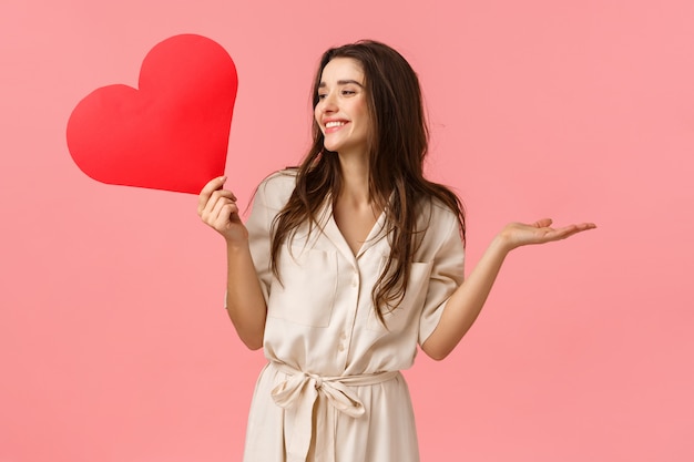 Foto surpresas, conceito de relacionamento e romance. atraente e despreocupada e feliz sorridente fêmea morena de vestido, levantando as mãos divertidas e alegres olhando para a esquerda, segurando o coração amor cartão, parede rosa