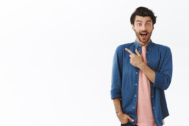 Surpreso atônito homem barbudo bonito na camiseta rosa apontando o canto superior esquerdo sorrindo animado boca aberta fascinado emocionado com a festa de natal convidando amigos promo de checkout