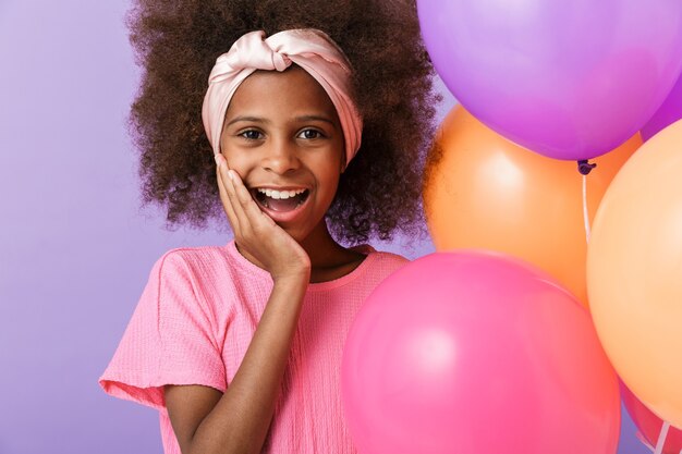 Surpreso chocado feliz jovem garota africana posando com balões isolados sobre a parede roxa.