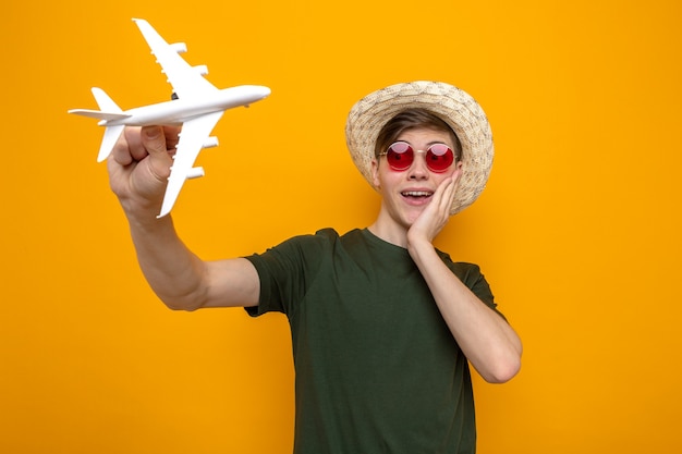 Surpreso, colocando a mão na bochecha, jovem bonito, usando um chapéu com óculos, segurando um avião de brinquedo isolado na parede laranja