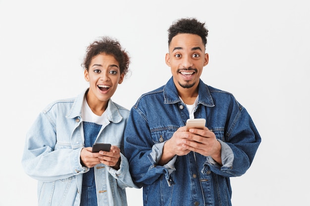 Surpreso feliz casal Africano em camisas jeans, segurando seus smartphones sobre parede cinza