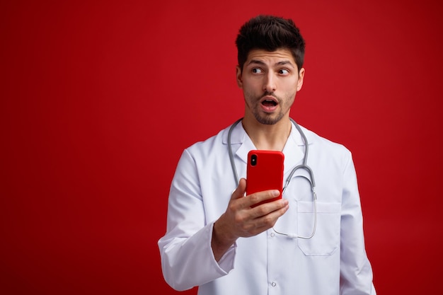 Surpreso jovem médico masculino vestindo uniforme médico e estetoscópio em volta do pescoço segurando o celular olhando para o lado isolado em fundo vermelho com espaço de cópia