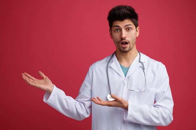 Surpreso jovem médico masculino vestindo uniforme médico e estetoscópio no pescoço, olhando para a câmera apontando para o lado com as mãos isoladas no fundo vermelho