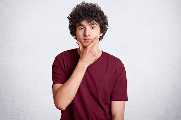 Surpreso macho encaracolado detém o queixo e olha com expressão chocada, tem olhos cheios de descrença, veste camiseta casual vermelha