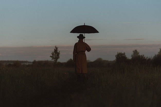 Surreale Silhouette eines Mannes in einem Anzug mit Regenschirm, der in einem Regenmantel auf einem Feld steht