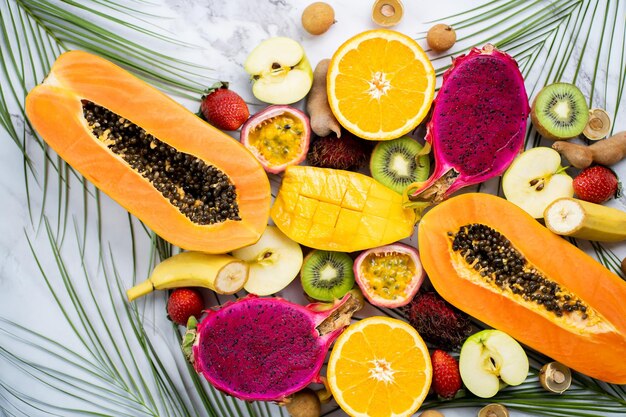 Surtido de diferentes frutas exóticas con hojas tropicales sobre fondo de mármol blanco. Vista superior, endecha plana. Alimentación saludable, dieta, frutas de temporada de verano.