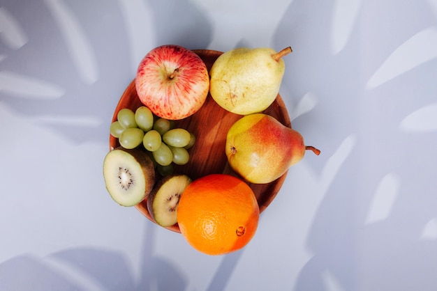 Surtido de frutas en un plato de madera y hojas sobre fondo blanco con sombra