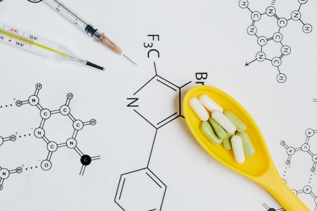 Surtido de píldoras y cápsulas en cuchara amarilla, jeringa, termómetro con fórmula química.