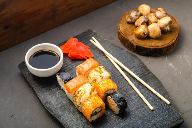 Surtido de rollos en una pizarra negra junto a palitos de salsa de soja y champiñones al horno sobre un fondo oscuro