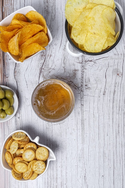 Foto surtido de snacks en casa con patatas fritas, cerveza, galletas saladas, aceitunas verdes y negras sobre mesa de madera