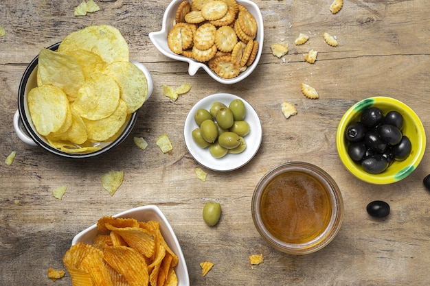 Surtido de snacks en casa con patatas fritas, cerveza, galletas saladas, aceitunas verdes y negras sobre mesa de madera