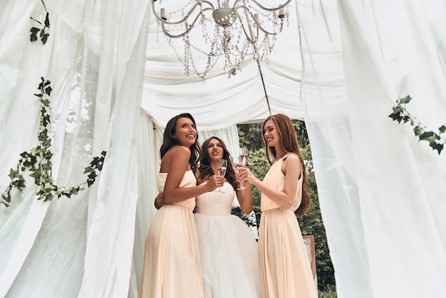 Con sus damas a su lado. Atractiva joven novia bebiendo champán con sus hermosas damas de honor mientras está de pie al aire libre