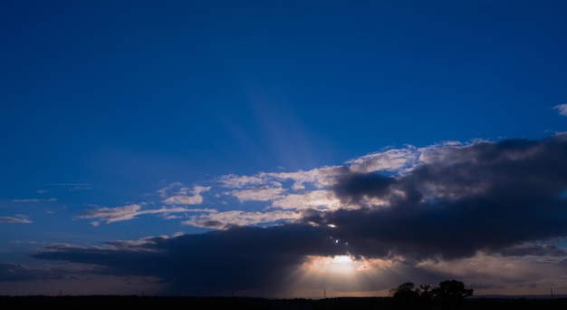 Suset-Himmellandschaft, Sonnenlicht scheint durch Wolkengebilde auf blauem Himmelshintergrund, Horizont, schöner Sonnenuntergang mit Silhouette, Hochspannungs-Elektro-Tansport, Hochspannungs-Übertragungsmast gegen Sonnenuntergang