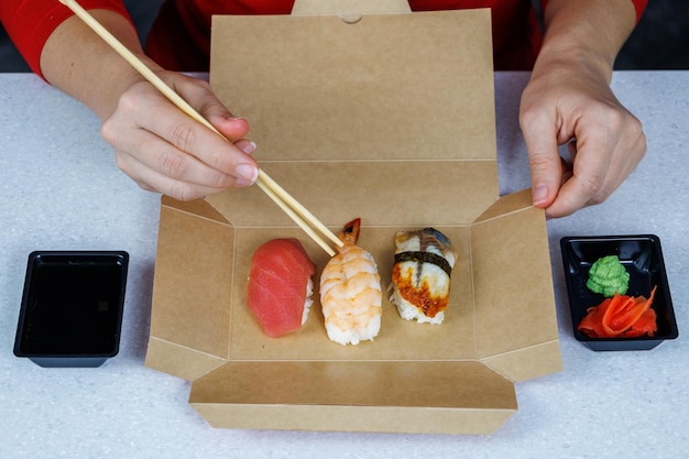 Sushi mit Thunfischgarnelen und Aal liegt in einem Karton auf dem Tisch in der Küche Das Mädchen im roten Pullover isst mit Essstäbchen Fast-Food-Konzept