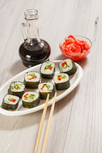 Sushi-Rollen mit Reis, Avocado-, Gurken-, Paprika- und Salatblättern auf Keramikplatte, Essstäbchen, Glasflasche mit Sojasauce und eingelegtem Ingwer in einer Schüssel. Vegetarisches Essen.