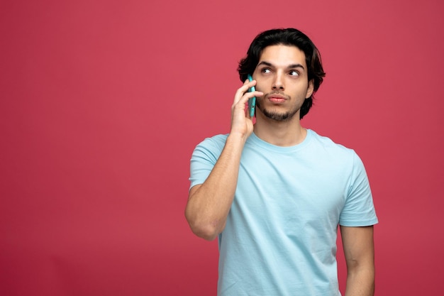 suspeito jovem bonito olhando para o lado enquanto fala no telefone isolado em fundo vermelho com espaço de cópia