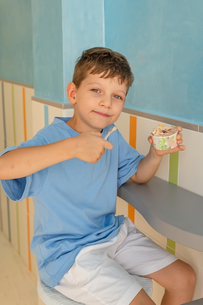 Sute menino de 6 anos em uma camiseta azul come sorvete com uma colher de um copo em uma gelateria em Roma Turismo tradicional de sobremesas com crianças