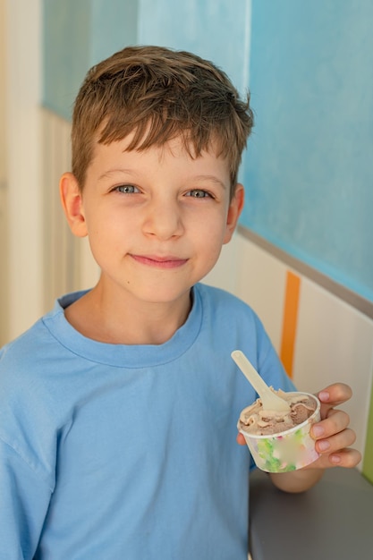 Sute menino de 6 anos em uma camiseta azul come sorvete com uma colher de um copo em uma gelateria em Roma Turismo tradicional de sobremesas com crianças