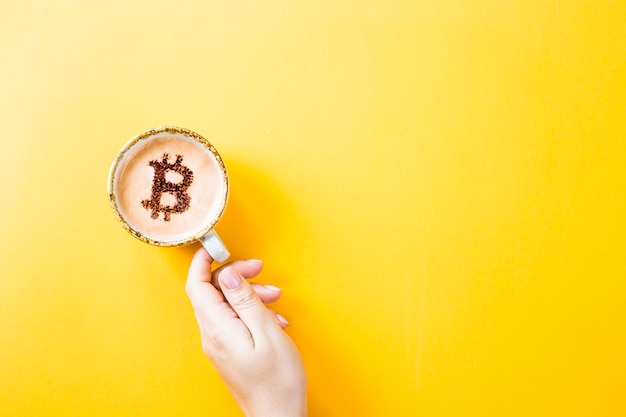 Symbol der Kryptowährung Bitcoin auf einem Tasse Kaffee auf einem gelben Hintergrund