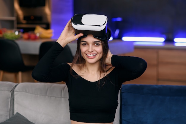 Sympathische, freudige, zufriedene Frau im Alter von 25 Jahren, die eine Augmented-Reality-Brille aufsetzt und in der modernen Wohnung an einem virtuellen Bildschirm arbeitet