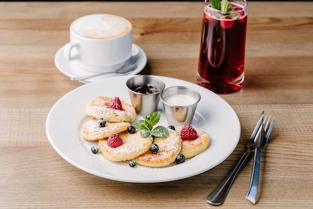 Syrniki con bayas en plato blanco aislado en mesa de madera en un café con crema agria y mermelada. Buñuelos de requesón. Limonada de café y bayas