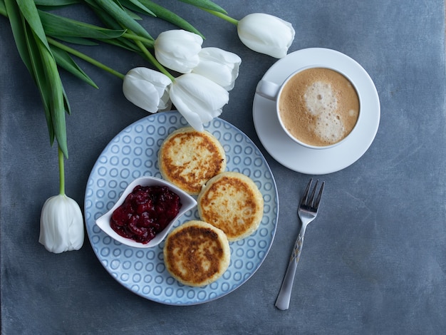 Syrniki-, Quark- oder Hüttenkäsepfannkuchen mit Kirschmarmelade und Kaffee auf grauem Hintergrund Mit weißen Tulpen, Draufsicht. Gesundes Diätfrühstück.