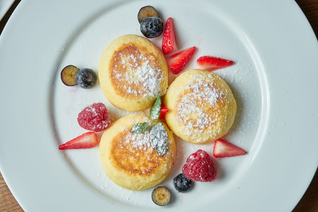 Syrniki ucraniano clásico con bayas, mermelada y crema agria en un plato blanco sobre una mesa de madera. Apetitoso postre de desayuno