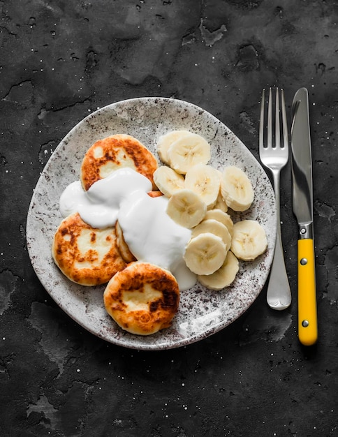 Syrnyky de queijo cottage com iogurte grego e banana em uma vista superior de fundo escuro