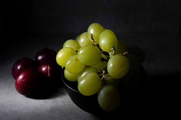 Szene im Darkfood-Stil mit einem Bündel grüner Weintrauben und einigen frischen roten Pflaumen