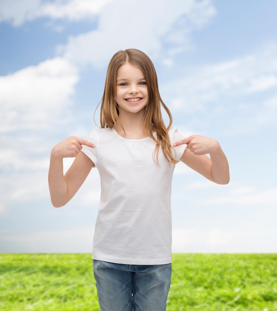 T-Shirt-Designkonzept - lächelndes kleines Mädchen im leeren weißen T-Shirt, das auf sich selbst zeigt