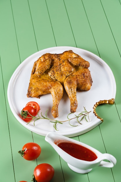 Tabaco de frango com molho de tomate, alecrim e tomate em um lindo prato branco sobre uma mesa verde. frango grelhado.