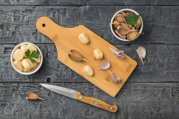 Una tabla de cortar, un cuchillo y tazones de ajo pelados y sin pelar sobre una mesa de madera. Una especia popular para la cocina.