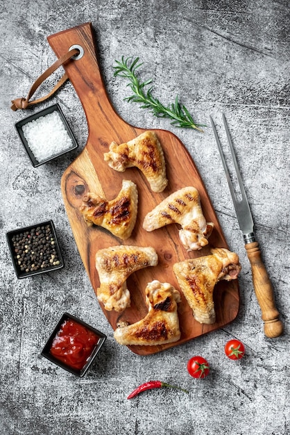 Una tabla de cortar de madera con alitas de pollo y un tazón pequeño de tomates y hierbas a un lado.