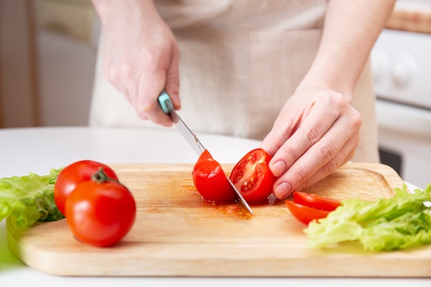 Tabla de cortar de madera con hierbas frescas, lechuga y verduras crudas sobre tablero de madera rústica. Fondo de cocina. Manos femeninas cortadas con un cuchillo Jugoso tomate rojo.