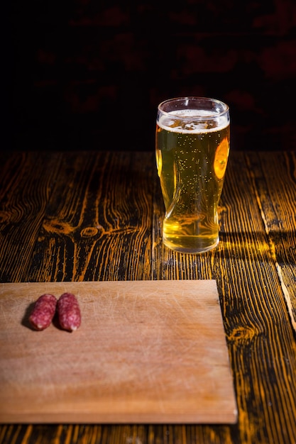 Tabla de cortar con salchichas en la mesa de madera cerca de un vaso lleno de cerveza