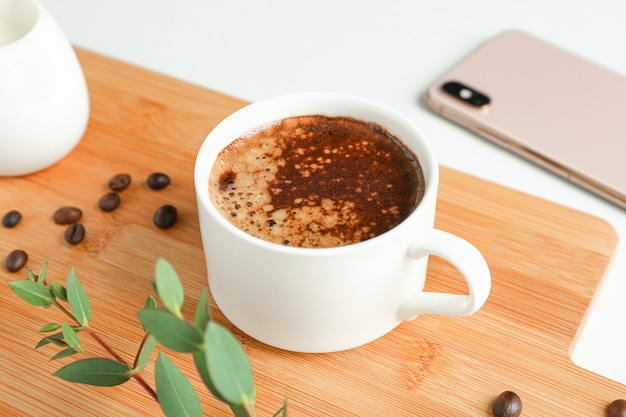 Tabla de cortar con una taza de café recién hecho, teléfono y rama de planta sobre fondo blanco.