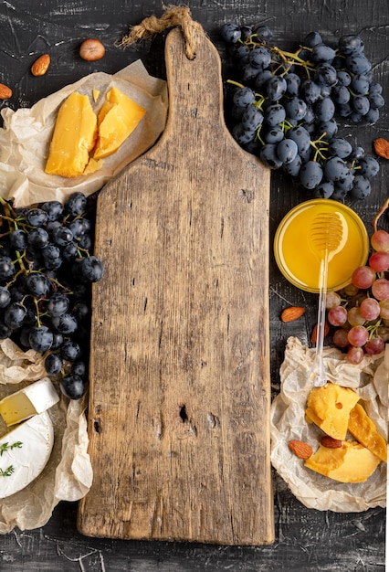 Tabla de cortar vintage de madera vacía en el centro del marco de miel, uvas, queso, bocadillos, otros ingredientes, bocadillos de gastronomía. Simulacros de espacio de copia o plantilla en tabla de cortar marrón. Vertical.