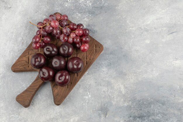 Tabla de madera de ciruelas rojas frescas y uvas sobre superficie de mármol.